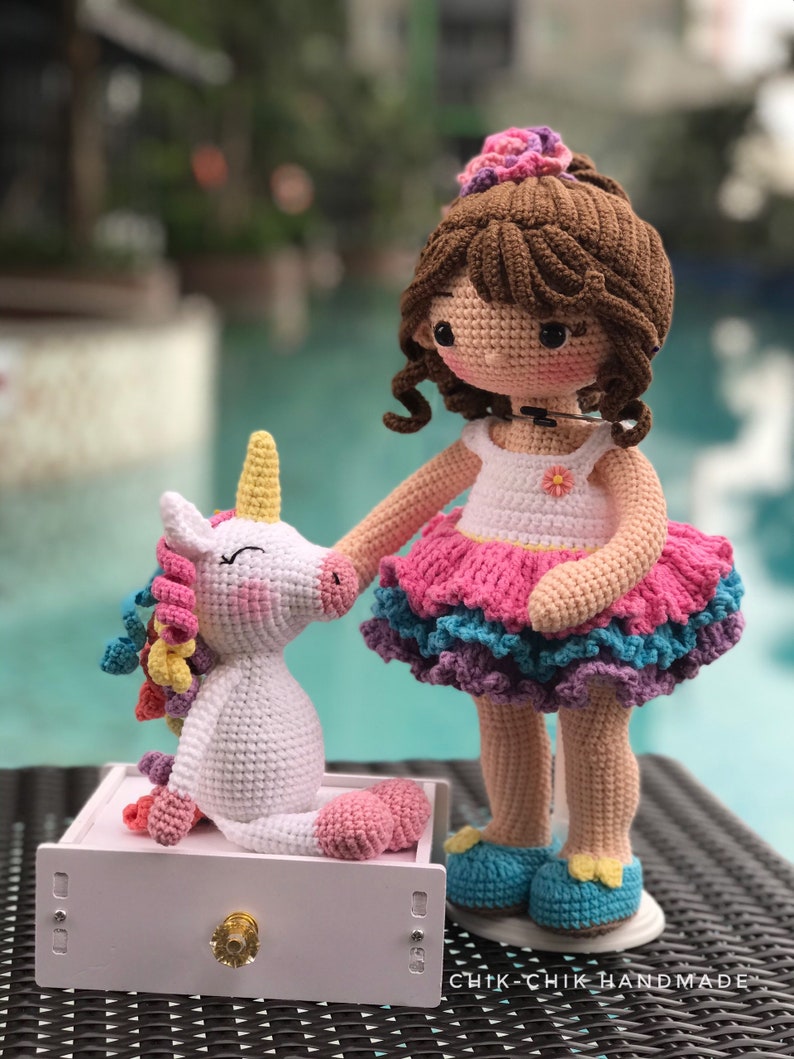 May include: A crocheted doll with brown hair and a pink and blue dress stands next to a white crocheted unicorn with a rainbow mane. The doll is wearing blue shoes and has a flower in her hair. The unicorn is sitting on a white drawer with a gold knob. The text "CHIK-CHIK HANDMADE" is visible on the bottom right corner of the image.