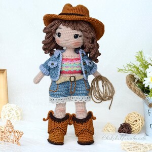 K&ouml;nnte beinhalten: H&auml;kelpuppe mit Jeansjacke, braunem Cowboyhut und blauen Jeans-Shorts. Die Puppe h&auml;lt ein braunes Seil in ihrer rechten Hand.