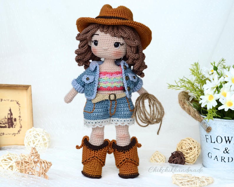 K&ouml;nnte beinhalten: H&auml;kelpuppe mit Jeansjacke, braunem Cowboyhut und blauen Jeans-Shorts. Die Puppe h&auml;lt ein braunes Seil in ihrer rechten Hand.