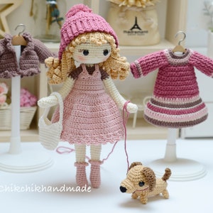 May include: A crocheted doll wearing a pink dress and a pink hat, holding a white crocheted bag and walking a brown crocheted dog. The doll is standing on a white base. There are two other crocheted outfits on display stands in the background.