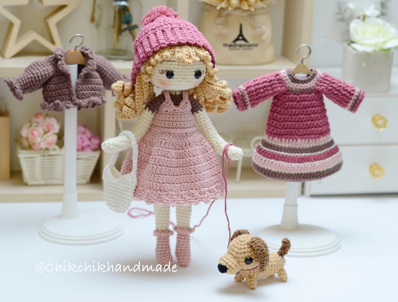 May include: A crocheted doll wearing a pink dress and a pink hat, holding a white crocheted bag and walking a brown crocheted dog. The doll is standing on a white base. There are two other crocheted outfits on display stands in the background.