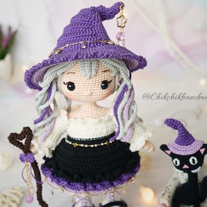 May include: A handcrafted crochet doll with light skin, grey hair, and a purple witch hat adorned with a gold chain and charm. The doll wears a white off-shoulder top and a black skirt with purple trim, holding a heart-topped staff near a black cat.