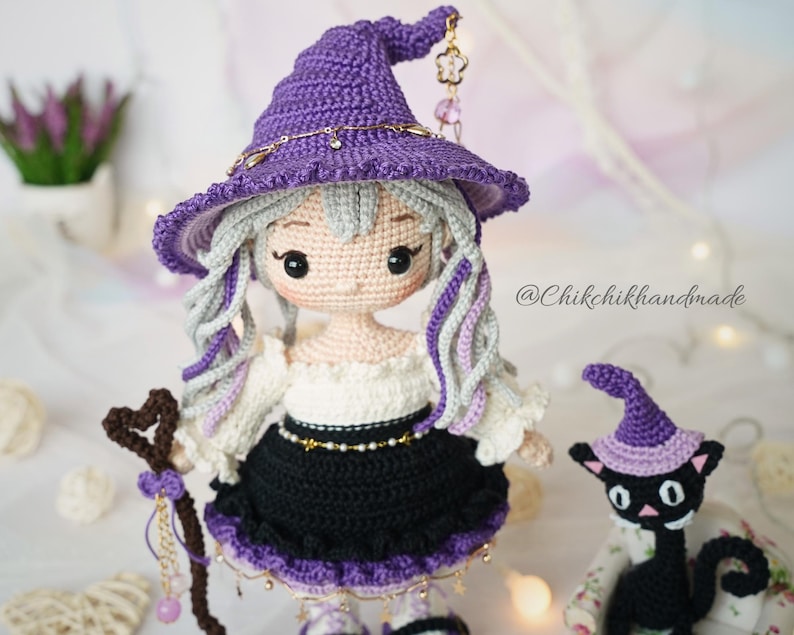 May include: A handcrafted crochet doll with light skin, grey hair, and a purple witch hat adorned with a gold chain and charm. The doll wears a white off-shoulder top and a black skirt with purple trim, holding a heart-topped staff near a black cat.