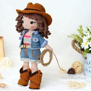 K&ouml;nnte beinhalten: H&auml;kelpuppe mit Jeansjacke, gestreiftem Shirt und braunem Cowboyhut. Die Puppe h&auml;lt ein Seil und tr&auml;gt braune Cowboystiefel.