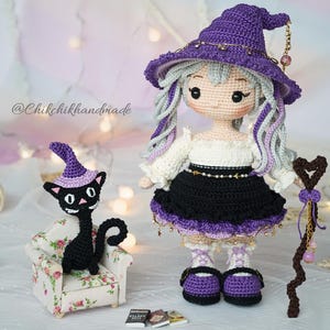 May include: A crochet doll wearing a purple witch hat, white blouse, and black skirt, holding a brown heart-topped staff. Beside the doll is a black crochet cat in a floral armchair, wearing a purple hat, with three miniature books on the floor.