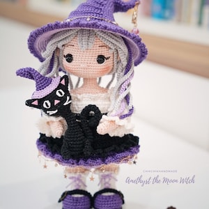 May include: A handcrafted crochet doll featuring a character with light gray hair wearing a purple witch hat and a black and white dress. The doll holds a small black cat wearing a purple hat. Text reads: CHIKCHIKHANDMADE Amethyst the Moon Witch.