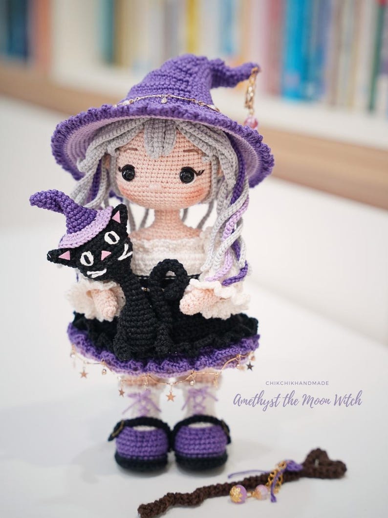 May include: A handcrafted crochet doll featuring a character with light gray hair wearing a purple witch hat and a black and white dress. The doll holds a small black cat wearing a purple hat. Text reads: CHIKCHIKHANDMADE Amethyst the Moon Witch.