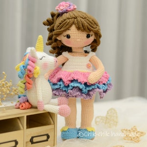 May include: A crocheted doll with brown hair wearing a white dress with a pink, blue, and purple layered skirt. The doll is holding a white crocheted unicorn with a rainbow mane.
