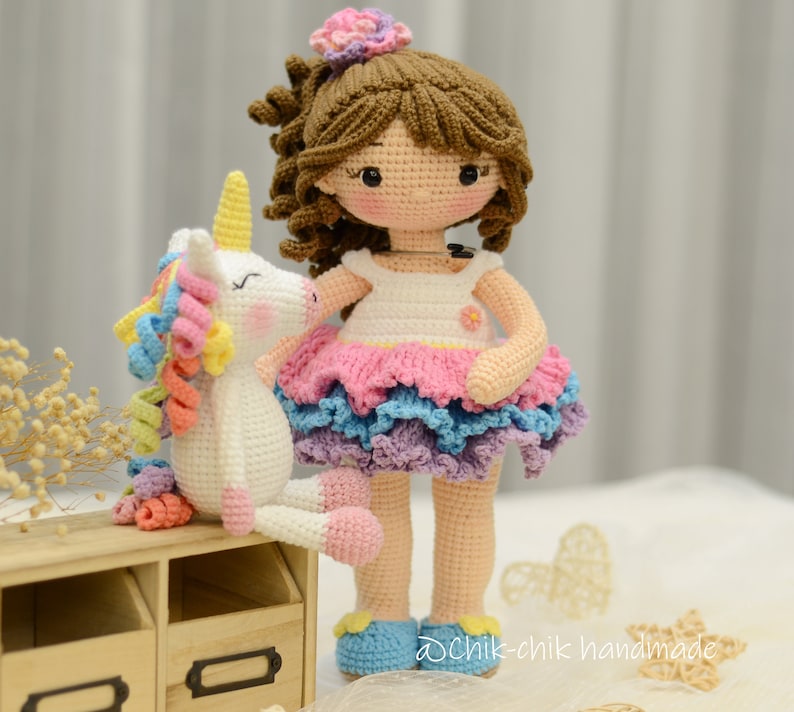 May include: A crocheted doll with brown hair wearing a white dress with a pink, blue, and purple layered skirt. The doll is holding a white crocheted unicorn with a rainbow mane.