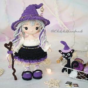 May include: A handcrafted crochet doll featuring light skin, long wavy silver hair, and a purple witch hat with gold charms. The doll wears a white blouse, black skirt, and purple shoes, holding a brown heart-topped staff. A small black cat wears a purple hat.