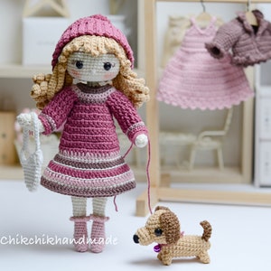 May include: A crocheted doll wearing a pink dress and a pink hat, holding a white crocheted bag. The doll is walking a brown and tan crocheted dachshund.
