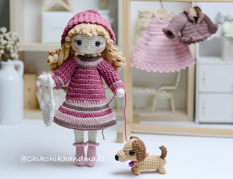 May include: A crocheted doll wearing a pink dress and a pink hat, holding a white crocheted bag. The doll is walking a brown and tan crocheted dachshund.