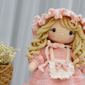 Pode incluir: Uma boneca de croch&ecirc; com roupa rosa e branca, um chap&eacute;u rosa e cabelo longo loiro. A boneca est&aacute; usando um avental branco com um la&ccedil;o rosa. A boneca &eacute; feita &agrave; m&atilde;o e o texto "Chnik-chnik handmade" &eacute; vis&iacute;vel no vestido da boneca.