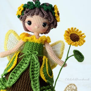 May include: A crocheted fairy doll with brown hair, wearing a green dress with yellow accents and a sunflower crown. The doll is holding a crocheted sunflower.
