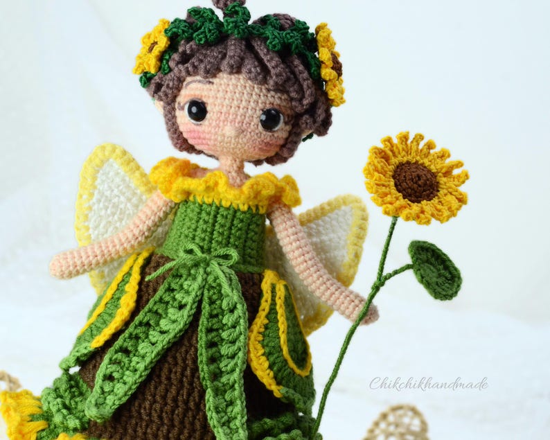 May include: A crocheted fairy doll with brown hair, wearing a green dress with yellow accents and a sunflower crown. The doll is holding a crocheted sunflower.