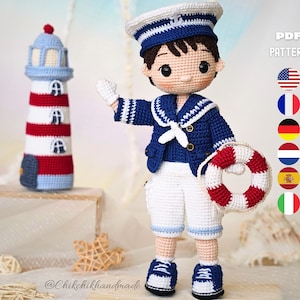 Puede incluir: Muñeco de marinero de ganchillo con chaqueta azul marino, pantalones cortos blancos y gorro de marinero, sosteniendo un salvavidas rojo y blanco. Un faro de ganchillo con rayas rojas y blancas y una parte superior azul está en el fondo. El muñeco mide unos 25 cm.