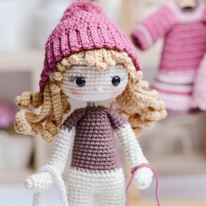 May include: A crocheted doll with blonde hair wearing a pink hat, a brown sweater, and white trousers. The doll is holding a white crocheted bag and a pink yarn.