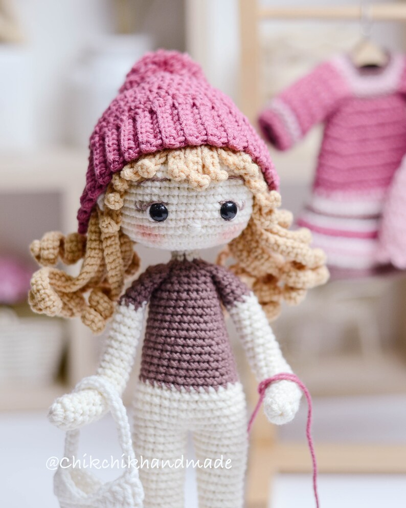 May include: A crocheted doll with blonde hair wearing a pink hat, a brown sweater, and white trousers. The doll is holding a white crocheted bag and a pink yarn.