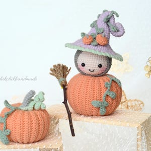 May include: A crocheted pumpkin with a smiling face wearing a purple and green witch hat. The pumpkin is holding a small broom and is sitting on a white surface. There is a smaller crocheted pumpkin with green leaves in the foreground.