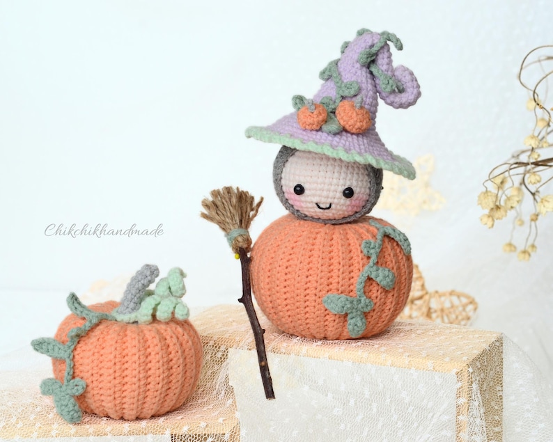 May include: A crocheted pumpkin with a smiling face wearing a purple and green witch hat. The pumpkin is holding a small broom and is sitting on a white surface. There is a smaller crocheted pumpkin with green leaves in the foreground.