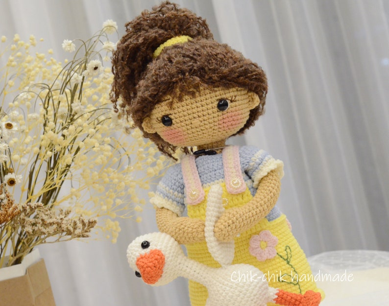 May include: A crocheted doll with brown curly hair wearing a yellow and blue outfit holds a white crocheted goose. The doll has a pink blush on its cheeks and black eyes. The doll is holding the goose in its arms. The doll is standing on a white surface. The text "Chik-chik handmade" is visible in the bottom right corner of the image.