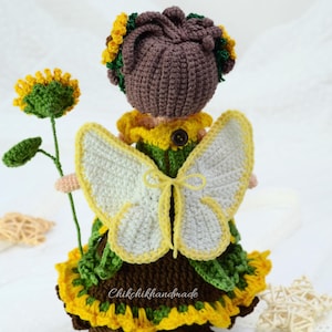 May include: Crocheted fairy doll with brown hair, a green and yellow dress, and white butterfly wings. The doll is holding a green sunflower stem.