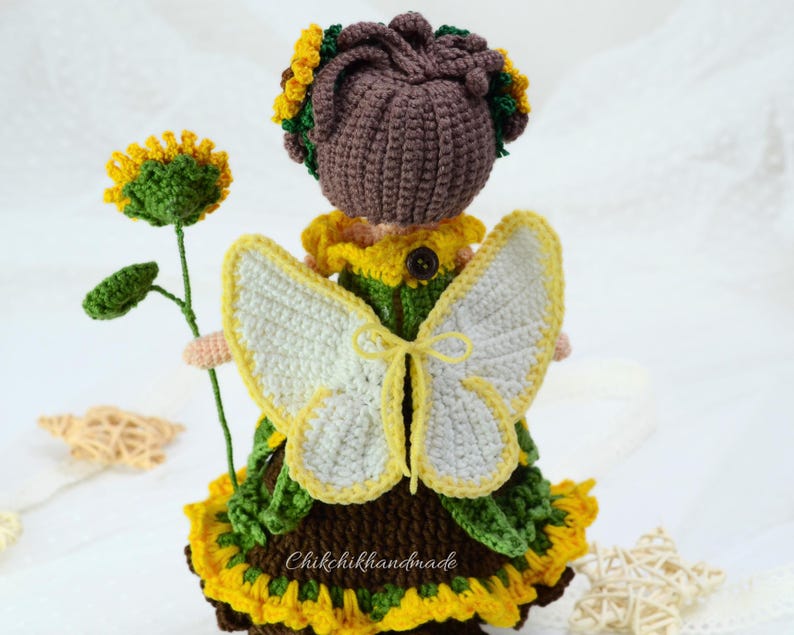 May include: Crocheted fairy doll with brown hair, a green and yellow dress, and white butterfly wings. The doll is holding a green sunflower stem.