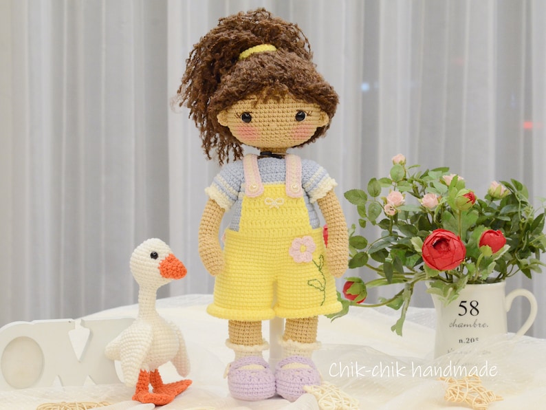 May include: A crocheted doll with brown hair in a ponytail wearing a yellow overall dress and a blue and white striped shirt. The doll is holding a small white crocheted goose.