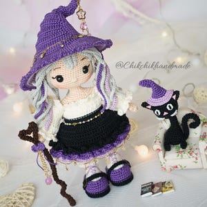 May include: A handcrafted crochet doll with light hair wearing a purple pointed hat, a white off-shoulder top, and a black skirt with purple trim. The doll holds a brown heart-topped staff. Beside it sits a small black crochet cat in a chair.