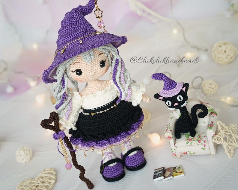 May include: A handcrafted crochet doll with light hair wearing a purple pointed hat, a white off-shoulder top, and a black skirt with purple trim. The doll holds a brown heart-topped staff. Beside it sits a small black crochet cat in a chair.