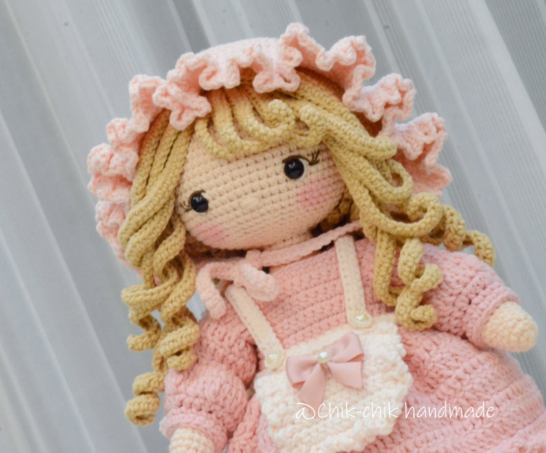 Pode incluir: Uma boneca de croch&ecirc; com roupas rosa e brancas, um chap&eacute;u rosa e cabelo longo loiro encaracolado. A boneca est&aacute; usando um avental branco com um la&ccedil;o rosa.