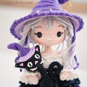 May include: A crochet doll named Amethyst the Moon Witch with light gray hair, wearing a purple pointed hat with a gold chain, and a white and black dress. The doll holds a small black cat wearing a matching purple hat with pink accents.