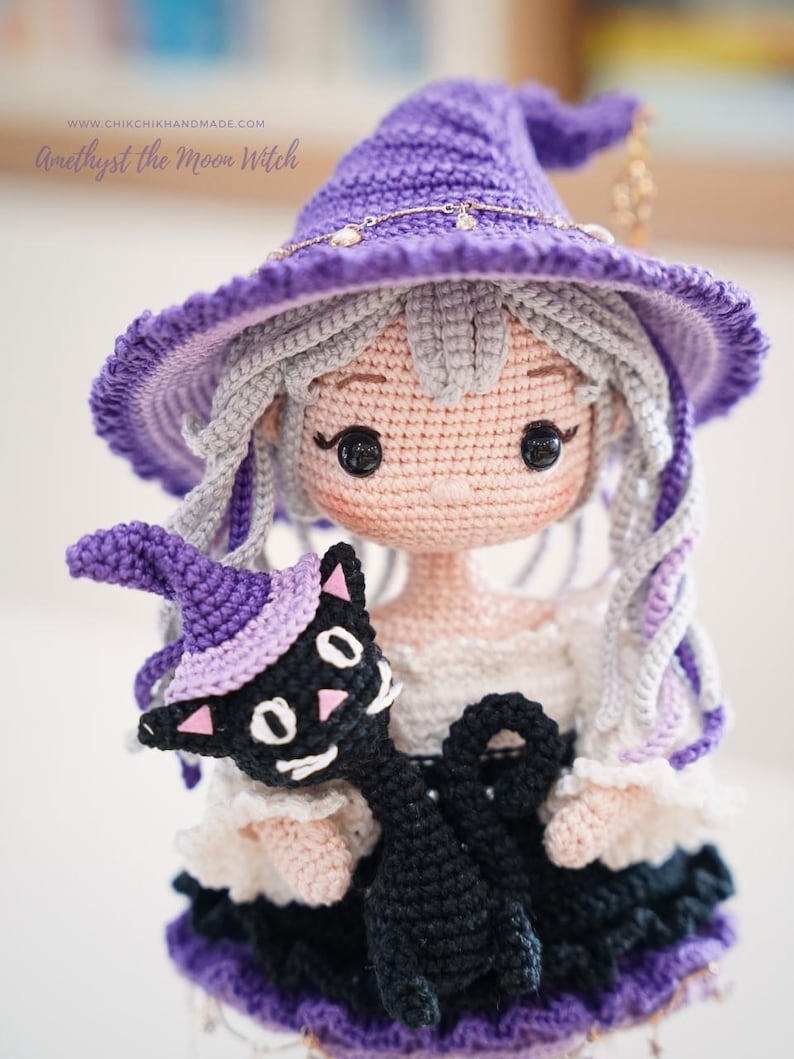 May include: A crochet doll named Amethyst the Moon Witch with light gray hair, wearing a purple pointed hat with a gold chain, and a white and black dress. The doll holds a small black cat wearing a matching purple hat with pink accents.