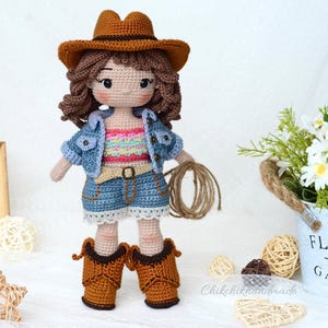 K&ouml;nnte beinhalten: H&auml;kelpuppe mit Jeansjacke, buntem Tanktop und braunem Cowboyhut. Die Puppe h&auml;lt ein Seil und tr&auml;gt braune Cowboystiefel.