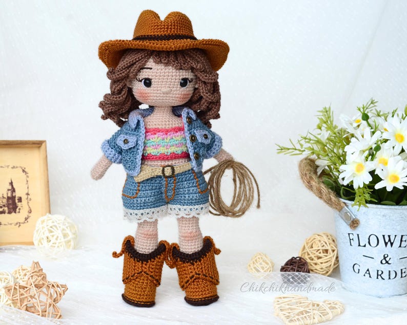 K&ouml;nnte beinhalten: H&auml;kelpuppe mit Jeansjacke, buntem Tanktop und braunem Cowboyhut. Die Puppe h&auml;lt ein Seil und tr&auml;gt braune Cowboystiefel.