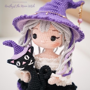 May include: A crocheted doll with light skin, grey hair, and a purple pointed hat, holding a small black cat wearing a matching purple hat. The text CHIKCHIKHANDMADE Amethyst the Moon Witch is visible above the doll's head in a script font.