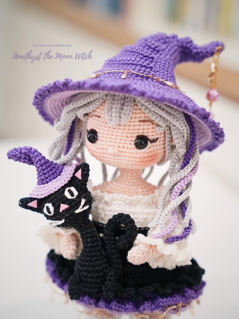 May include: A crocheted doll with light skin, grey hair, and a purple pointed hat, holding a small black cat wearing a matching purple hat. The text CHIKCHIKHANDMADE Amethyst the Moon Witch is visible above the doll's head in a script font.