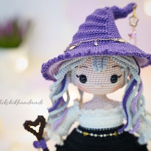 May include: A crocheted doll with light skin, black eyes, and long wavy hair in shades of light blue and purple. The doll wears a purple pointed hat with a gold chain and a charm, a white off-shoulder top, and a black skirt. Text reads @Chikchikhandmade.