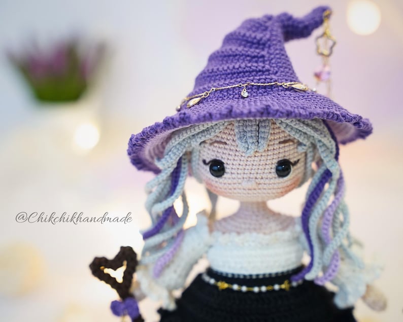 May include: A crocheted doll with light skin, black eyes, and long wavy hair in shades of light blue and purple. The doll wears a purple pointed hat with a gold chain and a charm, a white off-shoulder top, and a black skirt. Text reads @Chikchikhandmade.