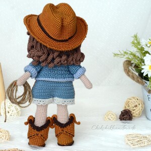 K&ouml;nnte beinhalten: H&auml;kelpuppe mit braunem Cowboyhut, blauem Jeanshemd und -shorts und braunen Cowboystiefeln. Die Puppe h&auml;lt ein Seil in ihren H&auml;nden.