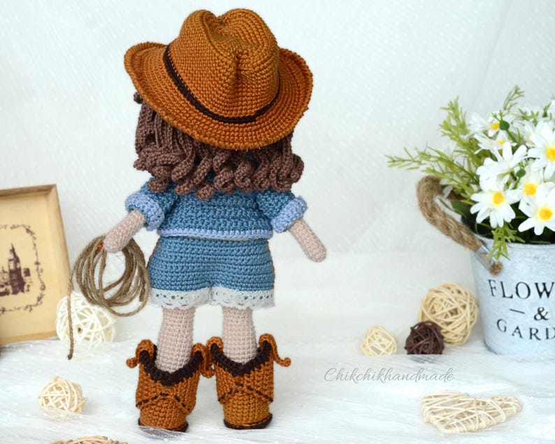 K&ouml;nnte beinhalten: H&auml;kelpuppe mit braunem Cowboyhut, blauem Jeanshemd und -shorts und braunen Cowboystiefeln. Die Puppe h&auml;lt ein Seil in ihren H&auml;nden.