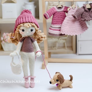 May include: A crocheted doll wearing a pink hat, brown top, white trousers, and pink shoes. The doll is holding a white crocheted bag and a lead attached to a brown and tan crocheted dachshund.