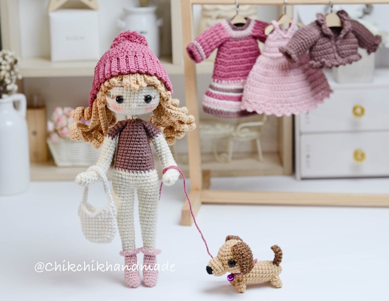 May include: A crocheted doll wearing a pink hat, brown top, white trousers, and pink shoes. The doll is holding a white crocheted bag and a lead attached to a brown and tan crocheted dachshund.