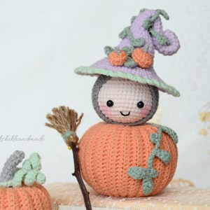May include: A crocheted pumpkin with a smiling face wearing a purple and green witch hat. The pumpkin is holding a small broom.