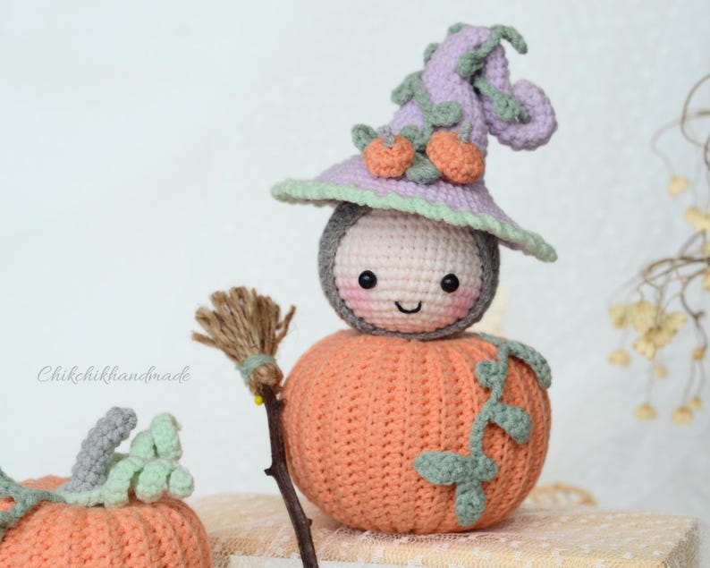 May include: A crocheted pumpkin with a smiling face wearing a purple and green witch hat. The pumpkin is holding a small broom.