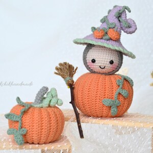 May include: A crocheted pumpkin witch doll with a purple hat and a broom. The doll is wearing a white face with a smiling mouth and black eyes. The doll is sitting on a wooden surface.