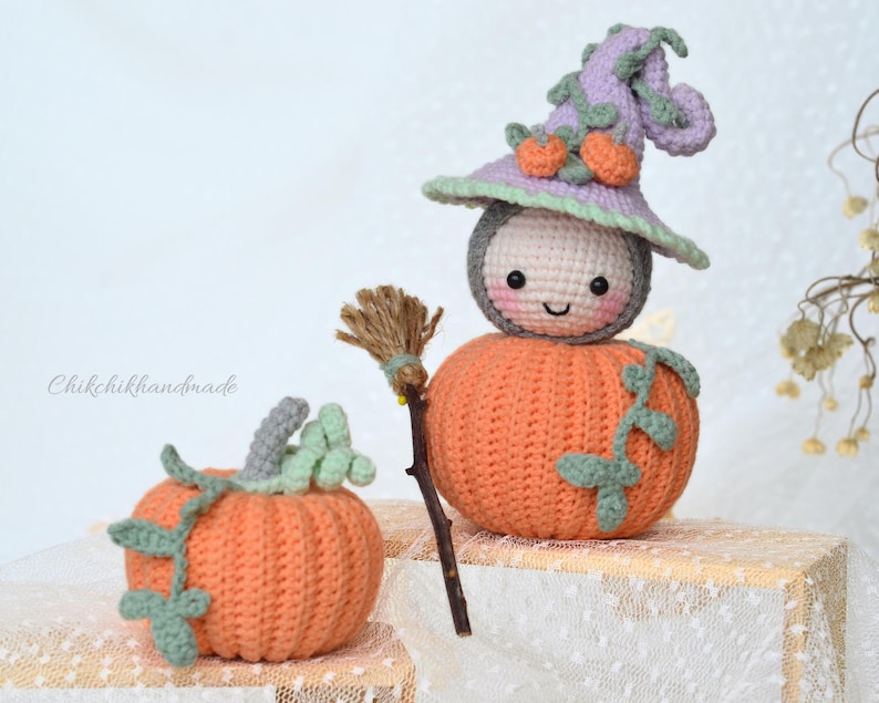 May include: A crocheted pumpkin witch doll with a purple hat and a broom. The doll is wearing a white face with a smiling mouth and black eyes. The doll is sitting on a wooden surface.