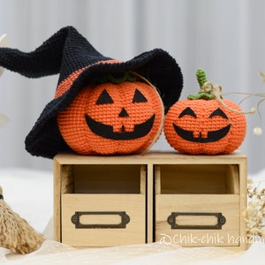 May include: Two crocheted pumpkins, one wearing a black witch hat, sitting on a wooden shelf with two drawers. The shelf has a label that says "@chik-chik handmade".