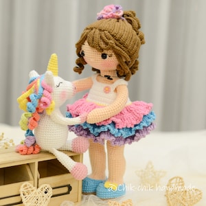 May include: A crocheted doll with brown hair wearing a pink and blue dress stands next to a crocheted unicorn with a rainbow mane. The doll is holding the unicorn's hoof. The doll is wearing blue shoes. The unicorn is sitting on a wooden shelf with two heart-shaped decorations.
