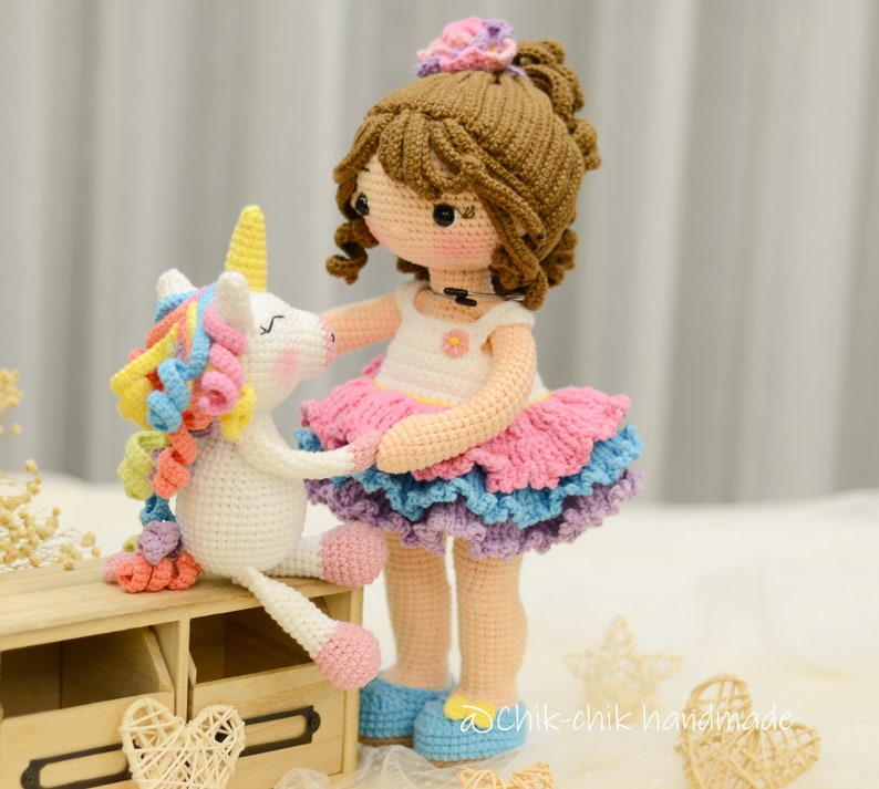 May include: A crocheted doll with brown hair wearing a pink and blue dress stands next to a crocheted unicorn with a rainbow mane. The doll is holding the unicorn's hoof. The doll is wearing blue shoes. The unicorn is sitting on a wooden shelf with two heart-shaped decorations.